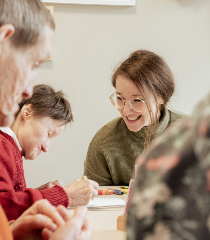 Menschen mit Behinderung sitzen an einem Tisch, eine Assistentin unterstützt.