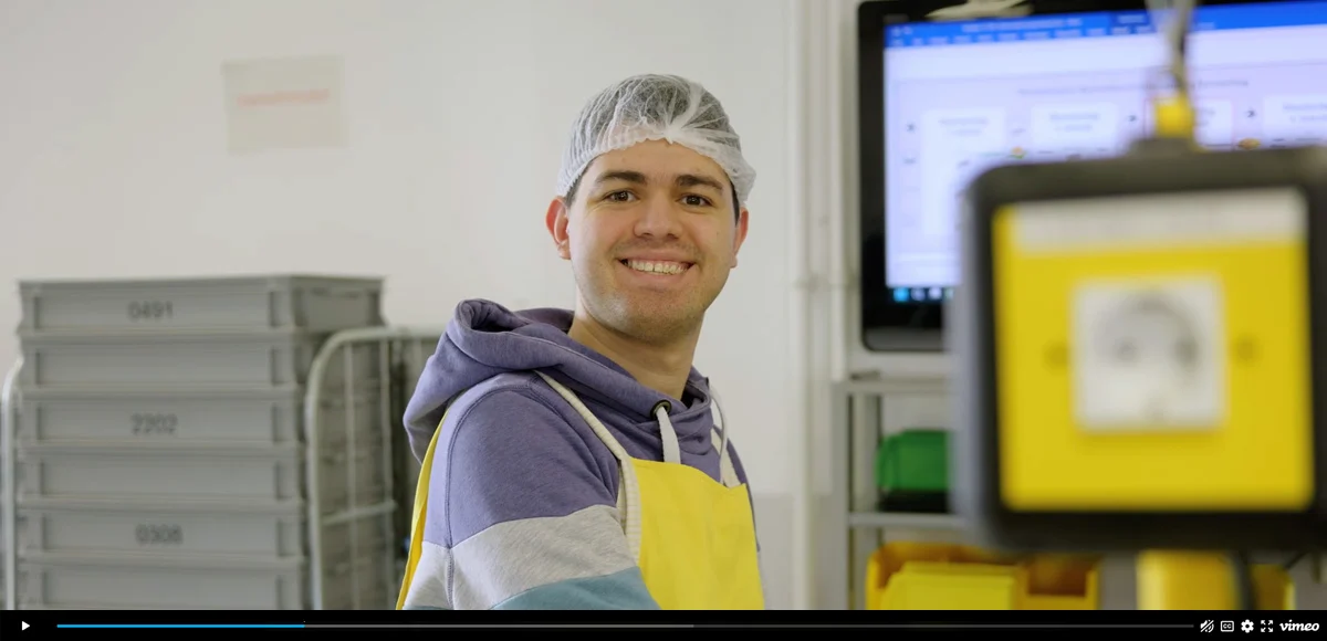 Mann steht in einem Produktionsraum, lächelt freundlich und trägt ein Haarnetz sowie eine Schürze.